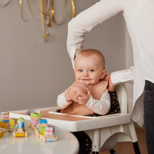 Ladda upp bild till gallerivisning, Ett litet barn som sitter i en barnstol och har en aprikos haklapp